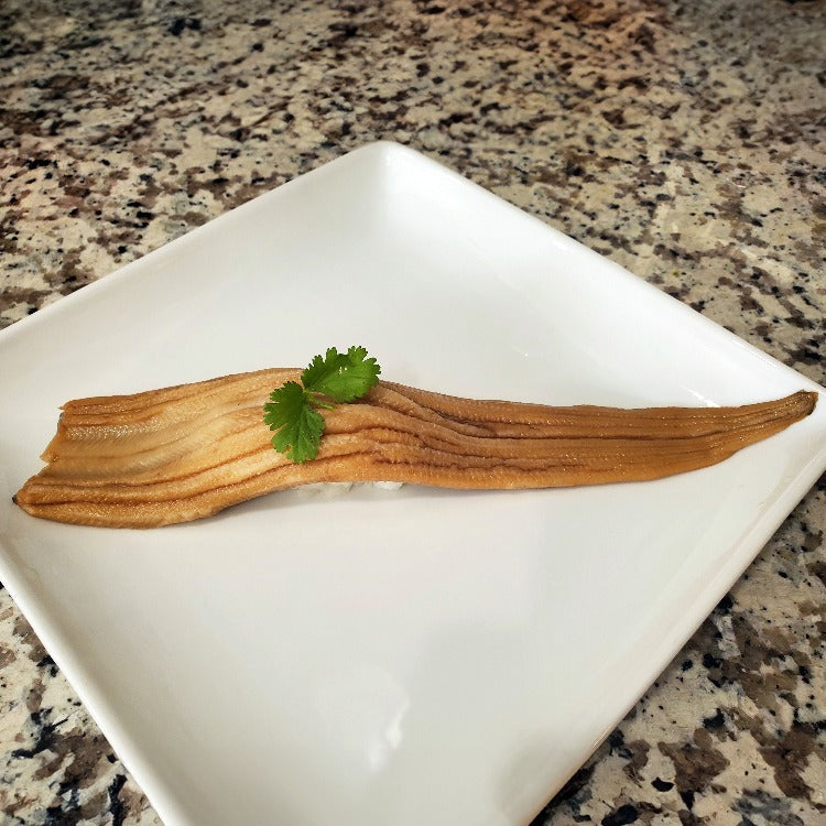 A single, cooked and seasoned eel fillet elegantly presented on a white square plate, garnished with a sprig of fresh parsley. The eel has a glossy, golden-brown exterior, indicative of a savory seasoning, and is prepared to emphasize its delicate texture and rich flavor