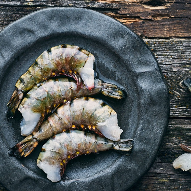 Four raw black tiger shrimp on a dark plate, showcasing their distinctive stripes and fresh appearance. The shrimp are ready for cooking, with their heads removed but shells intact