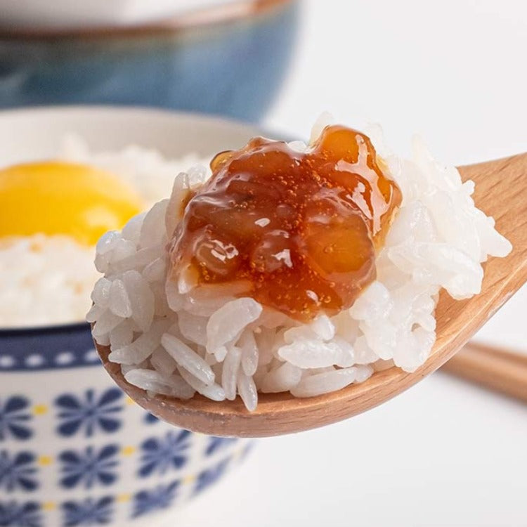 Wooden spoon holding a portion of white rice topped with Goremi seasoned shrimp with soy sauce, with a bowl containing rice and an egg yolk in the background. The shrimp is glistening and rich in sauce, ready to be mixed