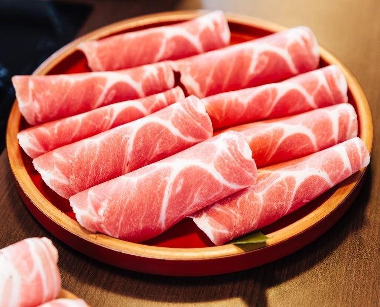 Rolled slices of pork shoulder presented on a round wooden plate, perfect for Korean BBQ or Japanese shabu-shabu. The rolls highlight the meat's even fat distribution and succulence