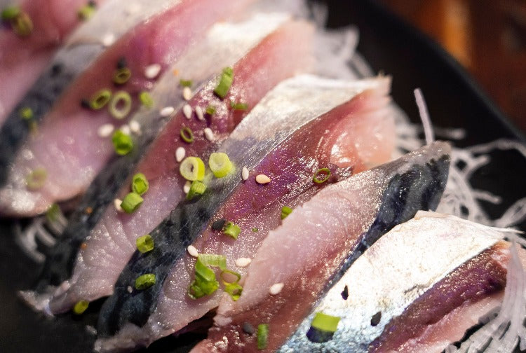 Sliced marinated mackerel garnished with green onions and sesame seeds on a dark ceramic plate