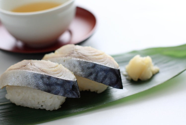 Two pieces of marinated mackerel sushi neatly presented on a bamboo leaf, accompanied by ginger and tea