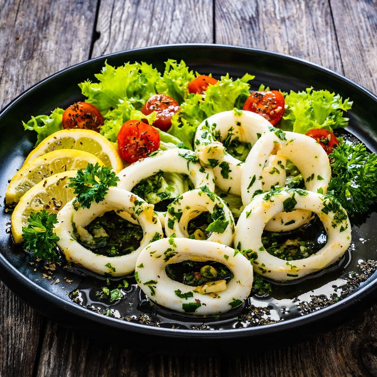 Grilled squid rings served on a black plate with a fresh salad, including cherry tomatoes, lettuce, and lemon slices, garnished with herbs