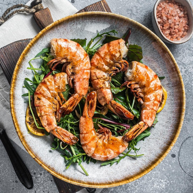 Plate of grilled black tiger shrimp served on a bed of fresh greens. The shrimp are beautifully charred and arranged in a circular pattern, highlighting their succulent texture and vibrant color