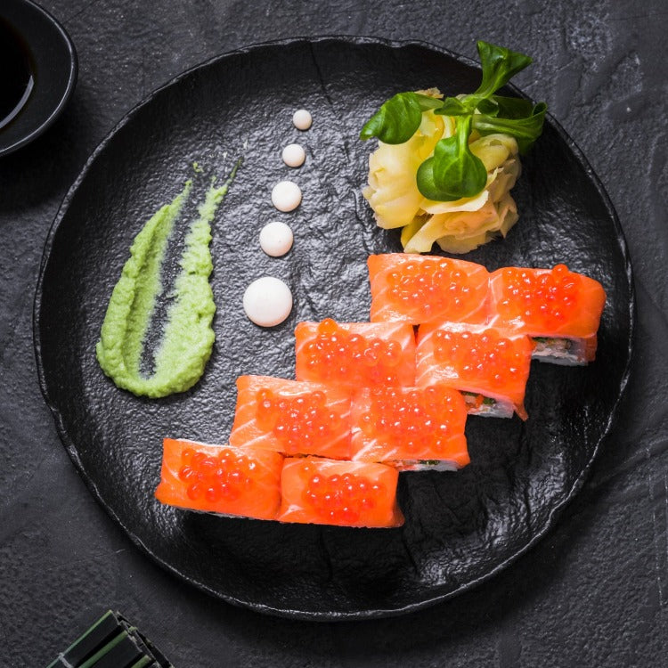 Gourmet sushi dish served with grated wasabi, sushi rolls topped with salmon roe, and garnished with fresh greens on a black plate.